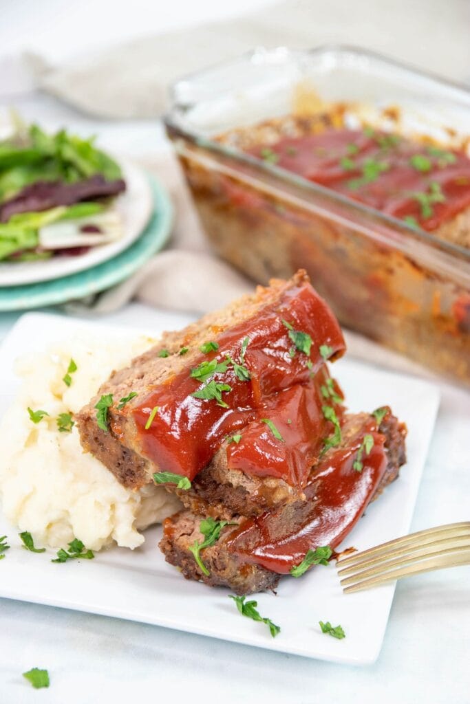 Finished crockpot meatloaf on plate with mashed potatoes and salad in background