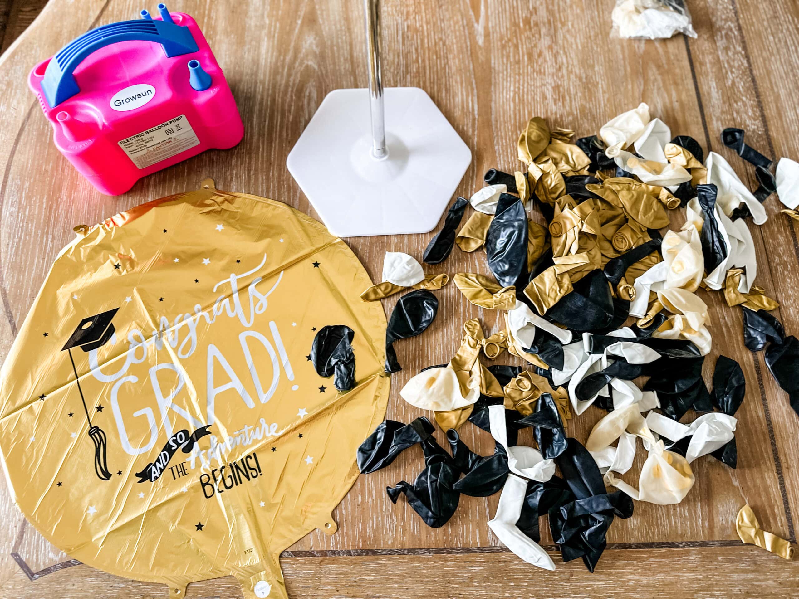 supplies for diy balloon bouquet on table - balloon pump, deflated balloons, large graduation mylar balloon, and balloon bouquet stand.
